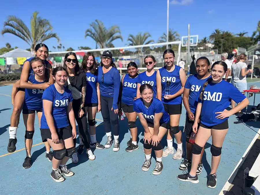 Group of young girls in blue volleyball uniforms with 'SMO' on shirts, posing on an outdoor basketball court after a game.