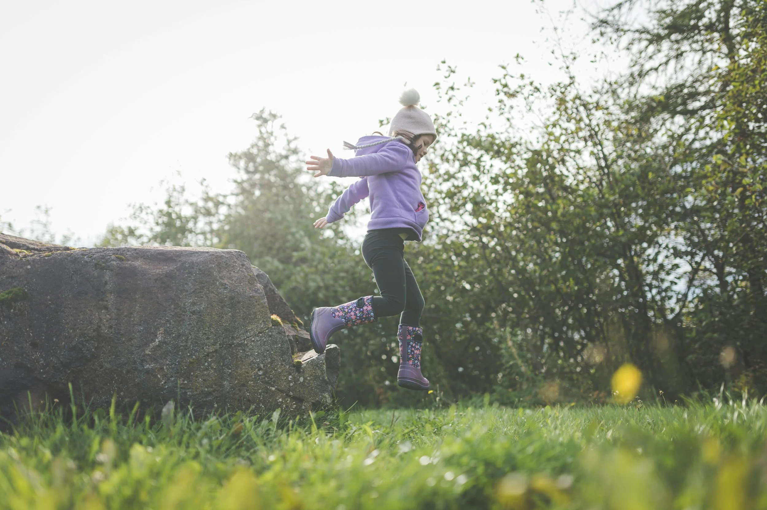 A young girl in a purple jacket, black pants, and floral rain boots is jumping off a rock onto grass in a park with green trees and bushes in the background.