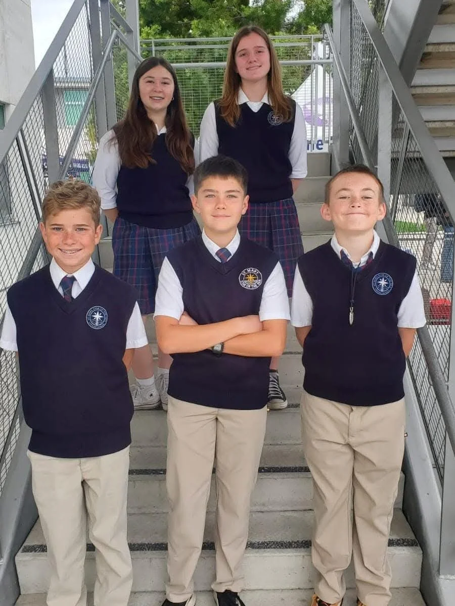 Six students in uniforms pose on outdoor staircase for a group photo. Four boys and two girls, all smiling, dressed in navy vests, white shirts, and khaki pants or skirts.