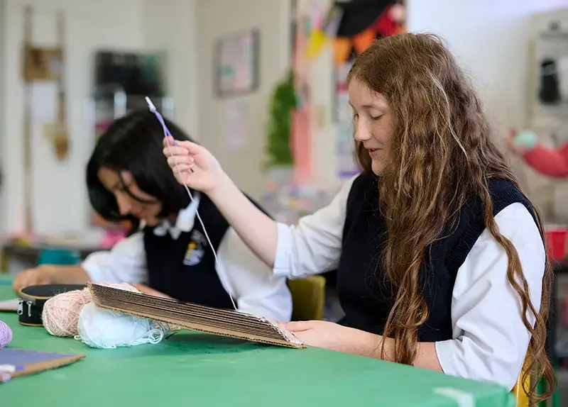 Two girls in school uniforms working on a craft project at a green table, with various supplies around them, in a classroom setting.