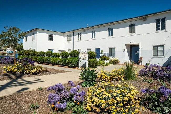 Exterior of a white two-story apartment complex with landscaped flower beds in the foreground, including purple and yellow flowers, green bushes, and an arched sign that reads 'S. Mesa Garden Apartments.'