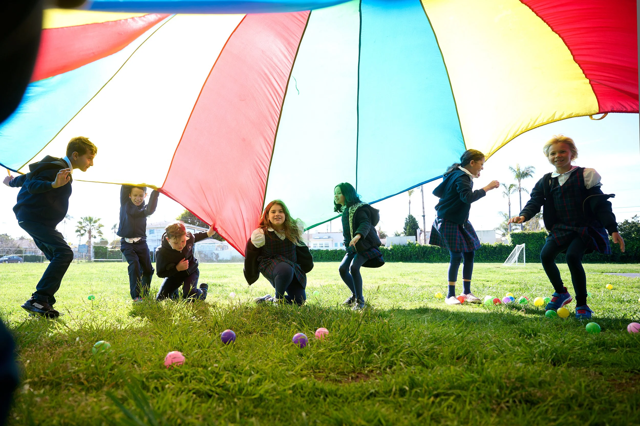 Children playing under a colorful parachute outdoors on a grassy field, with plastic eggs scattered on the ground and palm trees in the background.