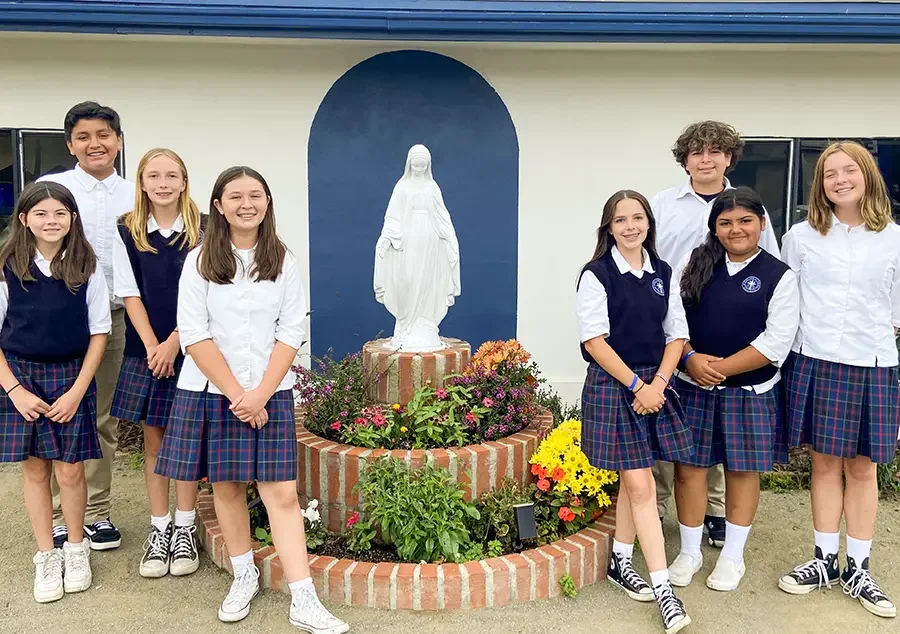 Group of students standing outdoors in front of a statue of the Virgin Mary surrounded by flowers.