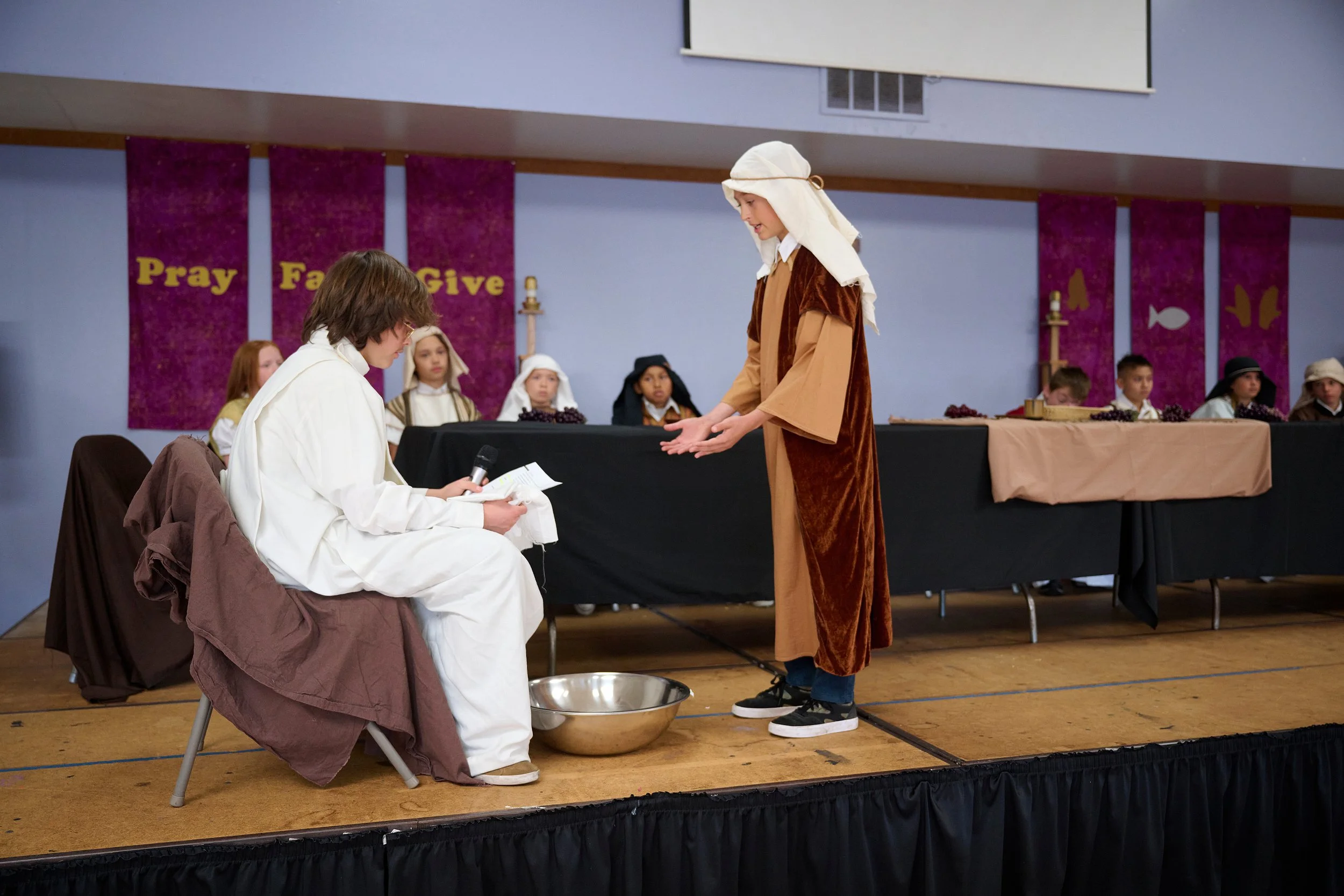 Children dressed in costumes performing a Nativity scene on a stage, with a purple banner reading 'Pray Faith Give' in the background.