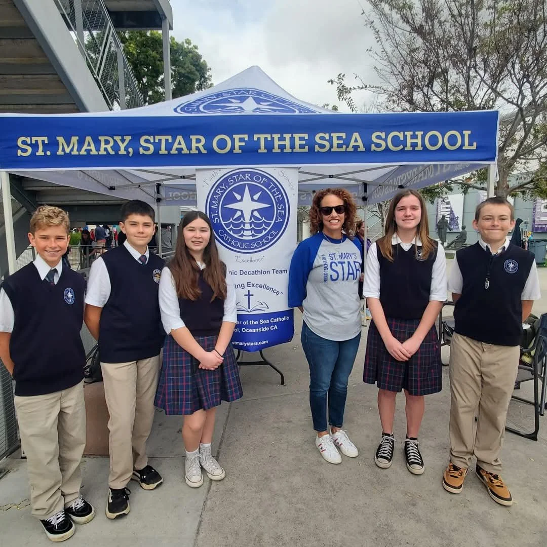 Group of students and adult woman standing in front of a tent with the sign 'St. Mary, Star of the Sea School.' The students are wearing school uniforms, and the woman is wearing a sweatshirt with the same school name.