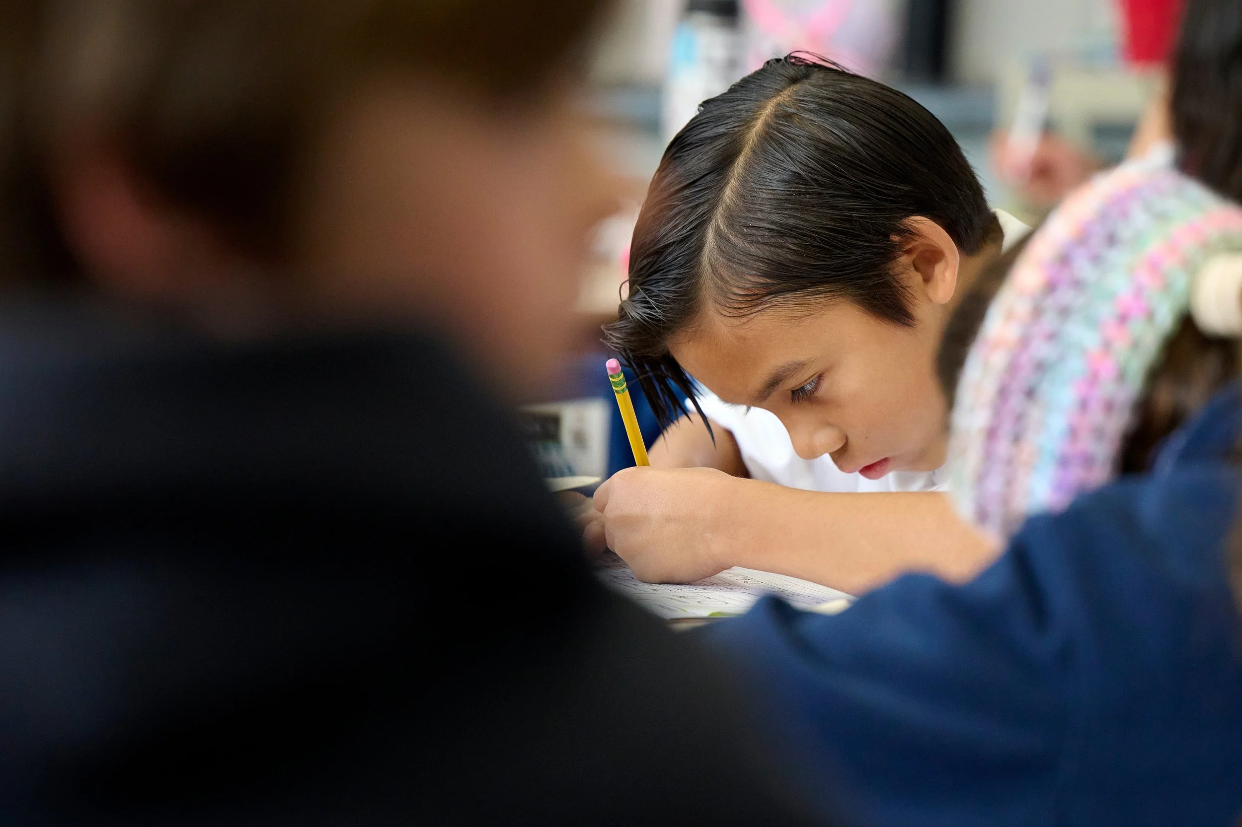 A young girl with dark, wet hair named with a middle part, diligently writing with a pencil in a classroom setting, surrounded by other students.
