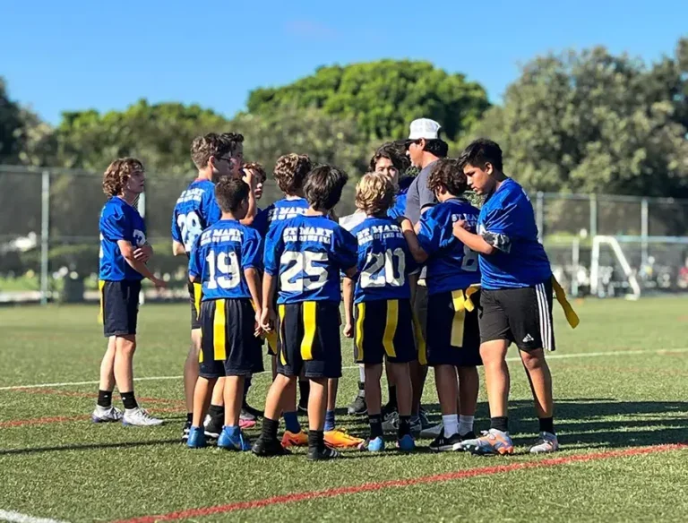 Youth football team in blue jerseys and black shorts huddling together on a grassy field with a coach during daytime.