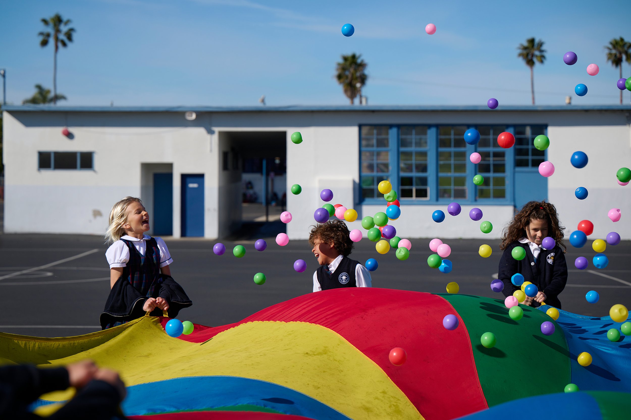 Three children playing with colorful balls outdoors near a school building with palm trees in the background.