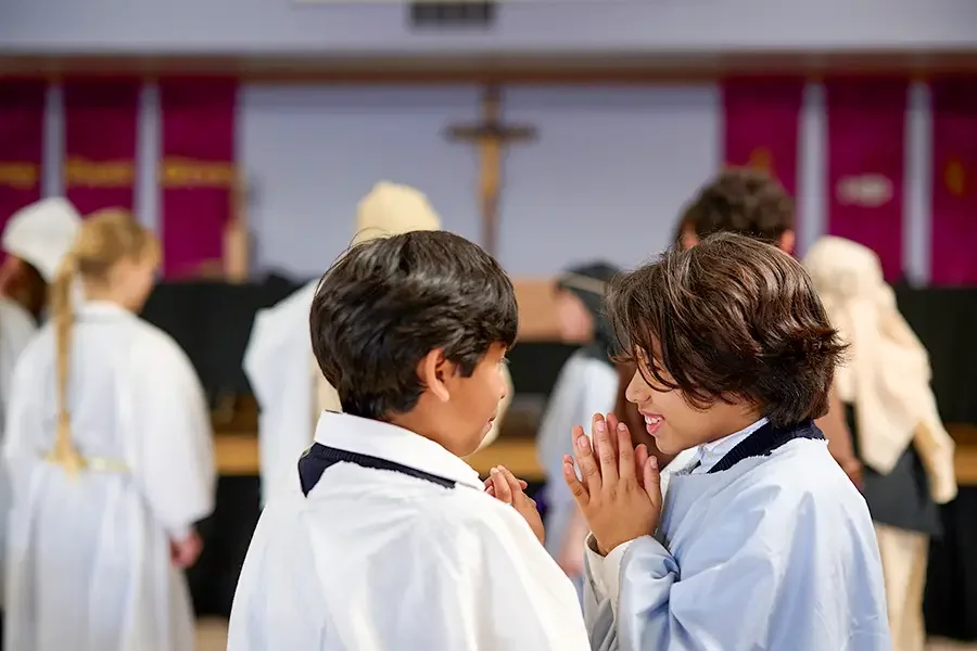 Two boys in white robes praying together inside a church, with other children in similar robes and a stained glass window in the background.