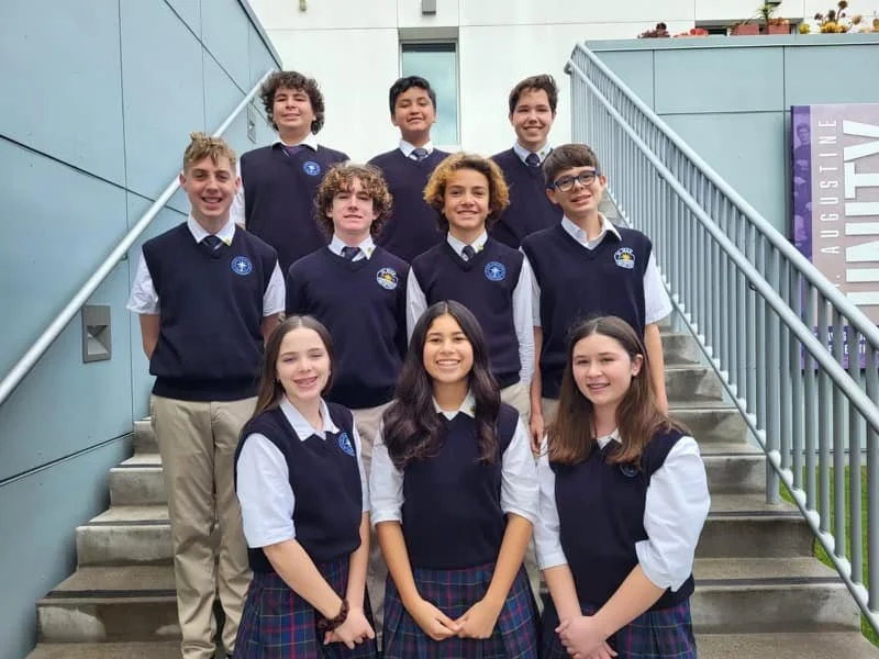 Group of ten students in school uniforms standing on outdoor stairs with a blue building wall on one side and a purple banner on the other.