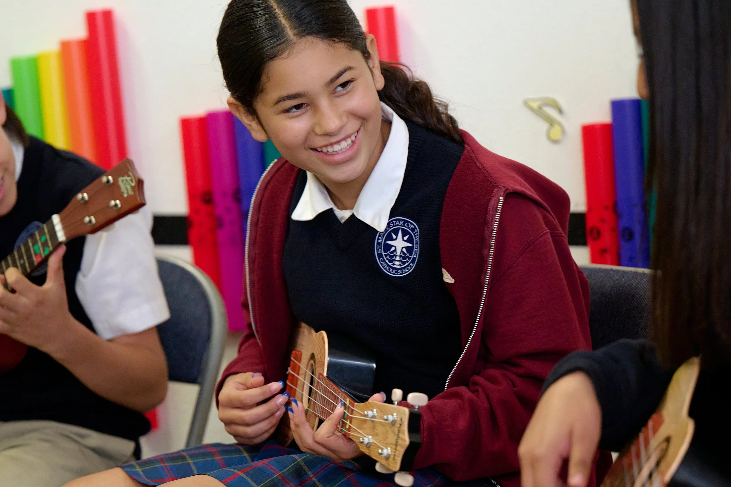 A girl smiling and sitting with a ukulele, wearing a school uniform with a dark sweatshirt and a red hoodie, in a classroom setting.