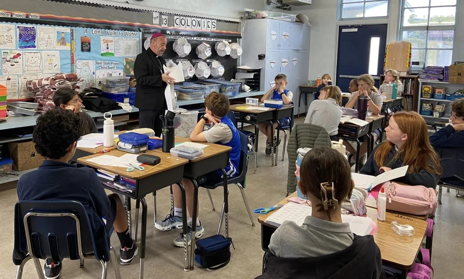 A classroom filled with elementary students sitting at their desks, listening to a teacher standing at the front. The teacher is holding and reading from a book while students look attentive. Classroom decorations, supplies, and a window are visible.