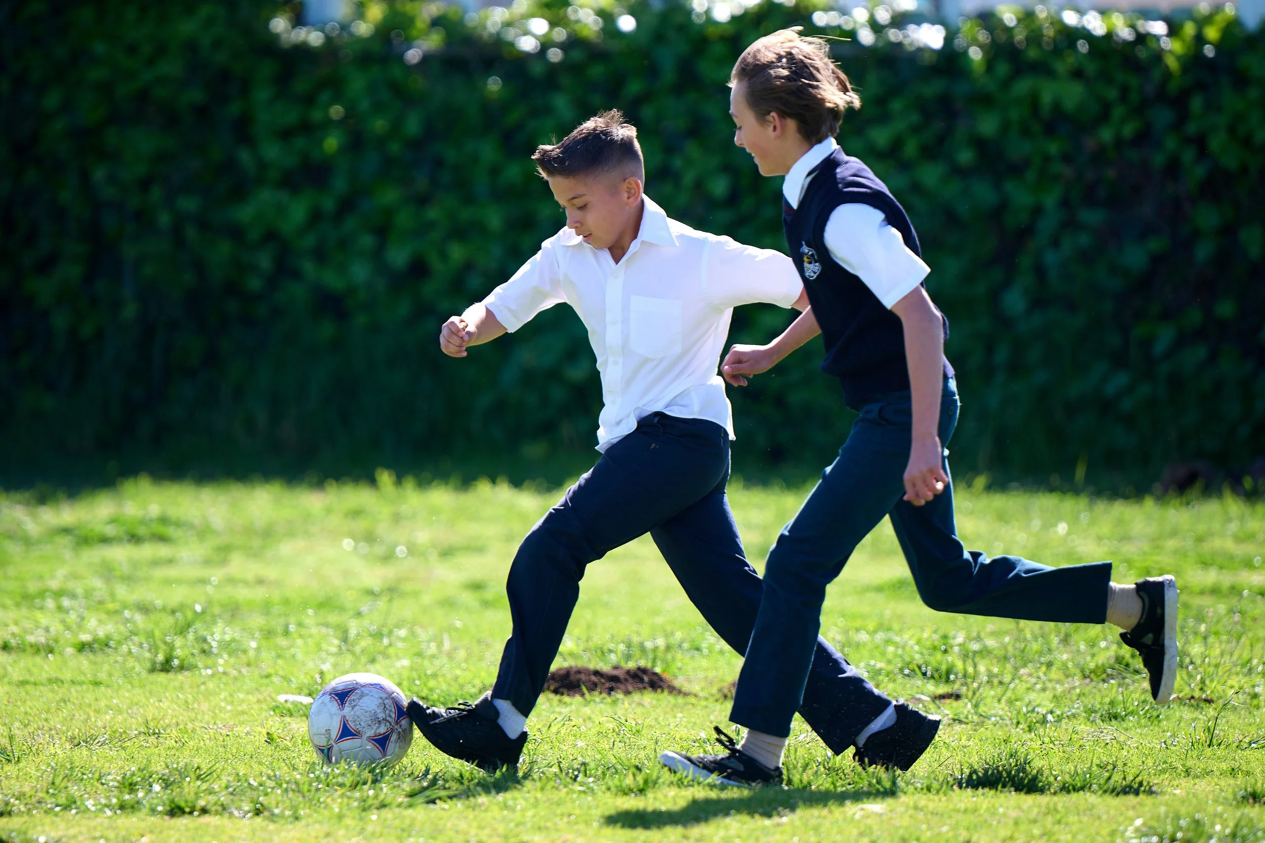Two boys in school uniforms playing soccer on a grassy field, one is kicking the ball while the other tries to block.
