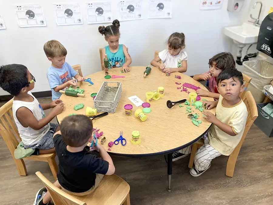 Children sitting around a round table engaging in arts and crafts with playdough and scissors in a classroom setting.
