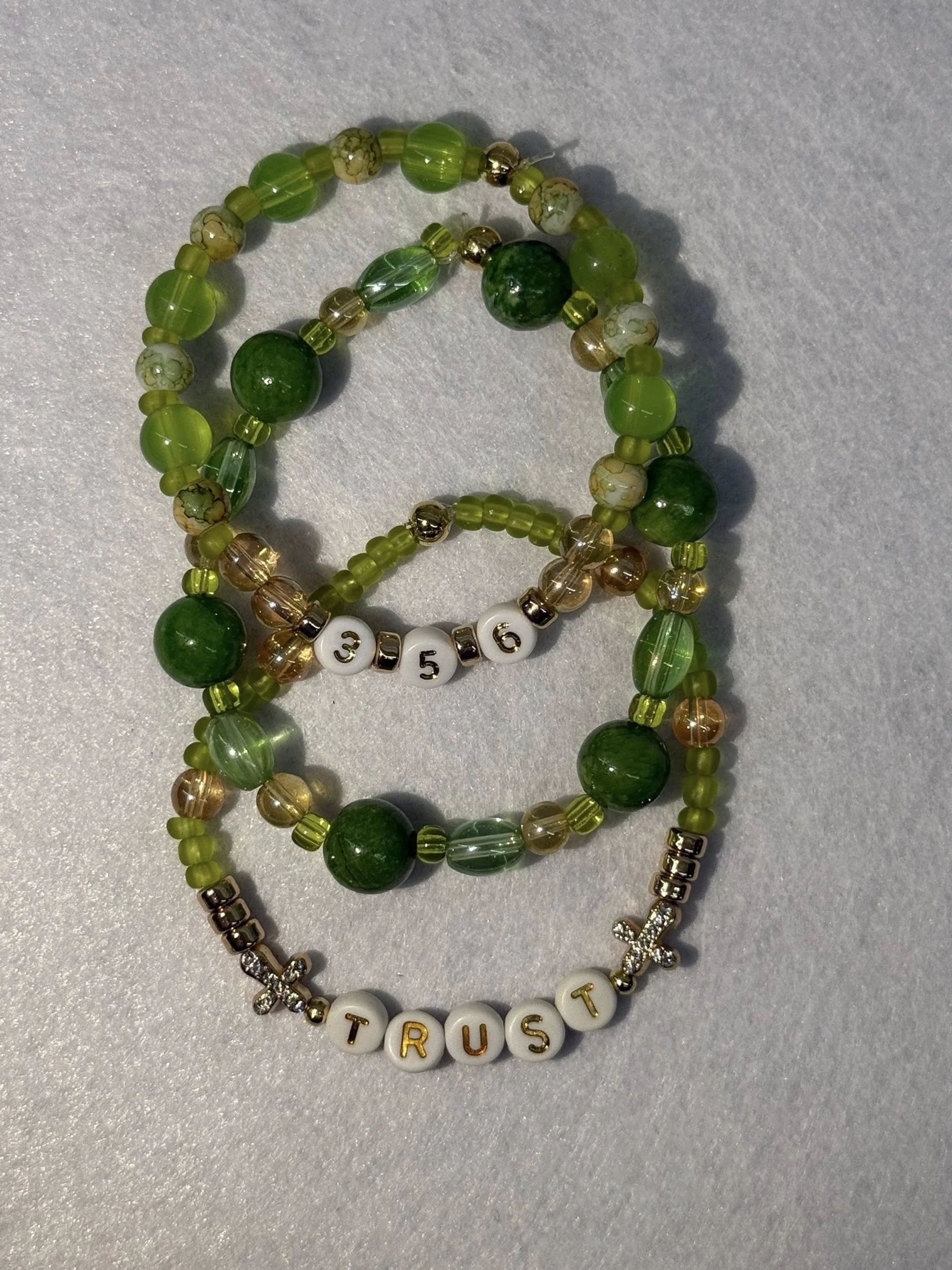 Three beaded bracelets, one with white beads spelling 'TRUST' and two with green beads, resting on a light gray surface.