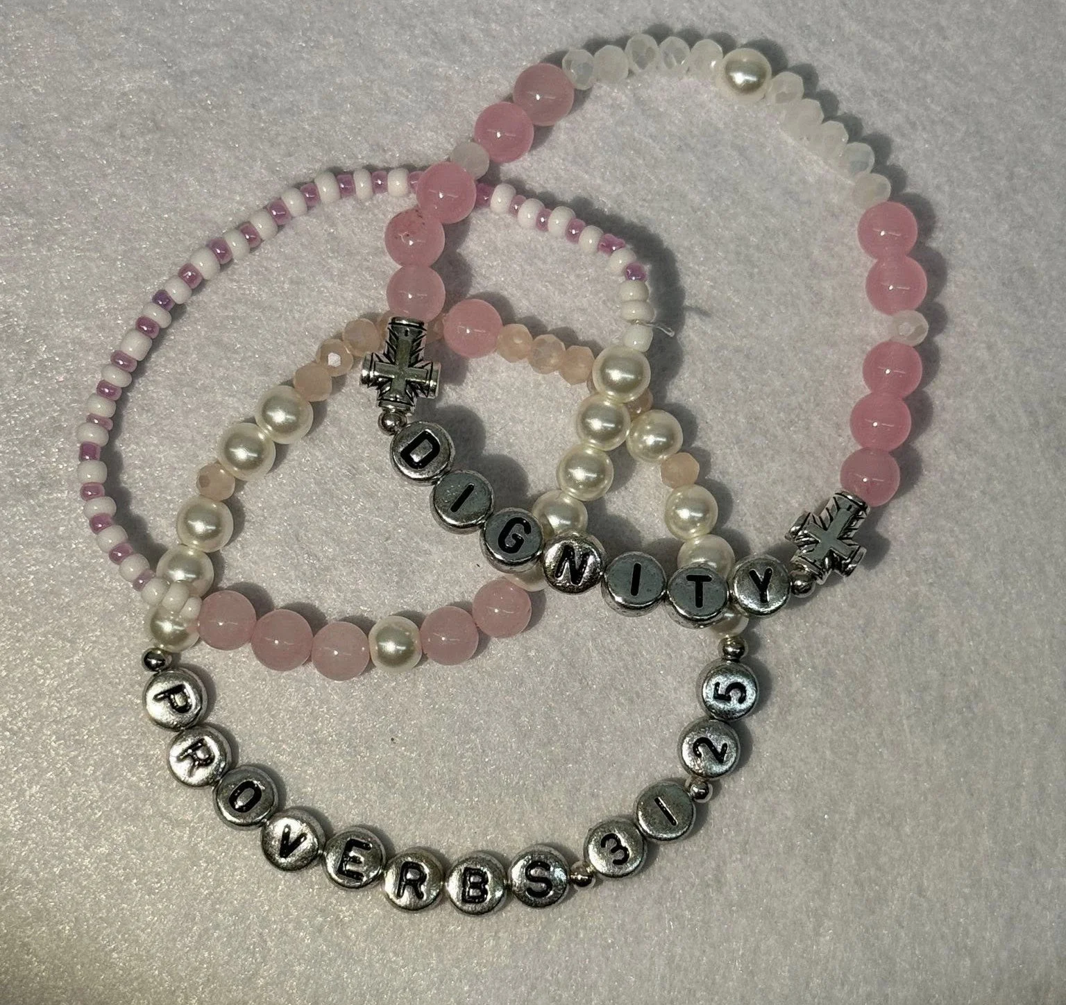 Three beaded bracelets with pastel pink, white, and clear beads. One bracelet spells 'LOYALTY' with alphabet beads, another spells 'LOVE BRB 13' with metallic letter beads, and the third has silver cross charms.