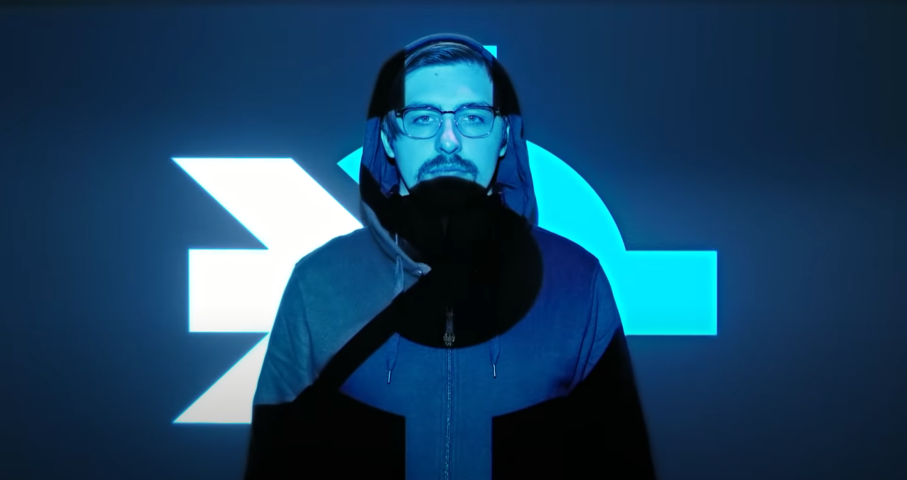 Young man with glasses and facial hair wearing a hoodie, standing in front of a blue digital background with geometric shapes.