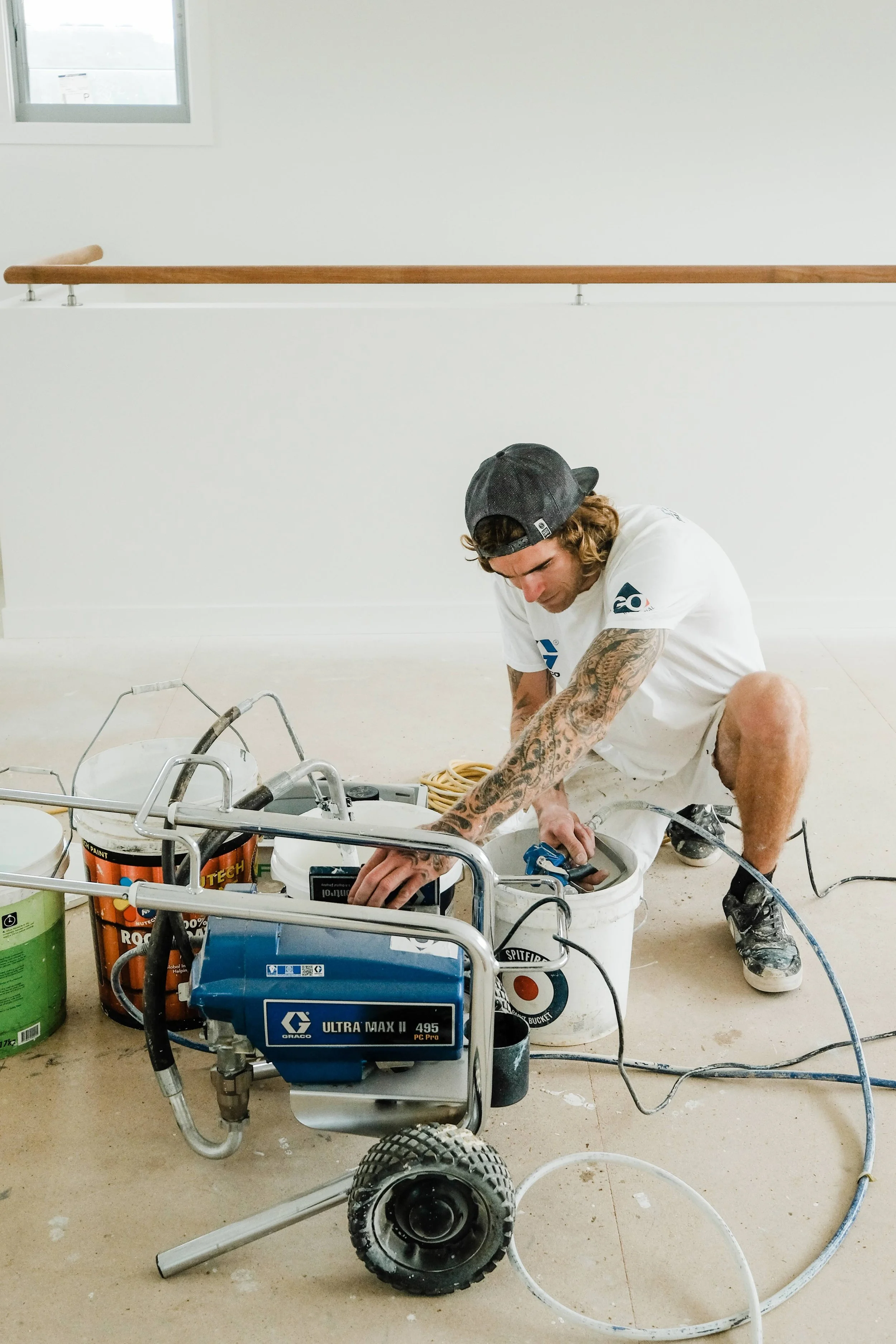 A man kneeling on the floor working with construction tools and equipment, surrounded by buckets of paint and paint sprayer, in a room with white walls and a small window.