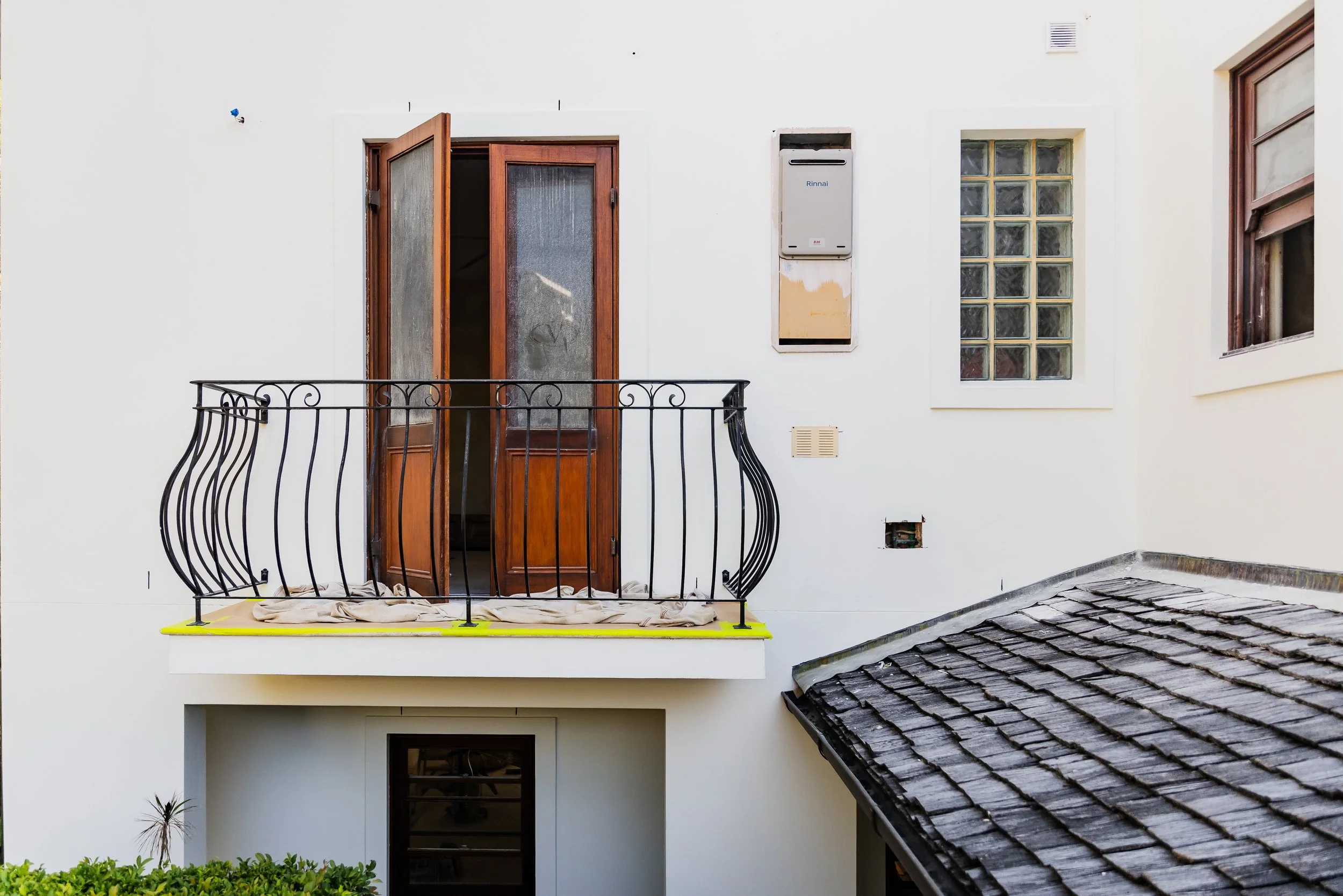A residential balcony with open wooden double doors, a decorative black metal railing, a white exterior wall with vents and windows, and a shingled roof over an adjacent building.