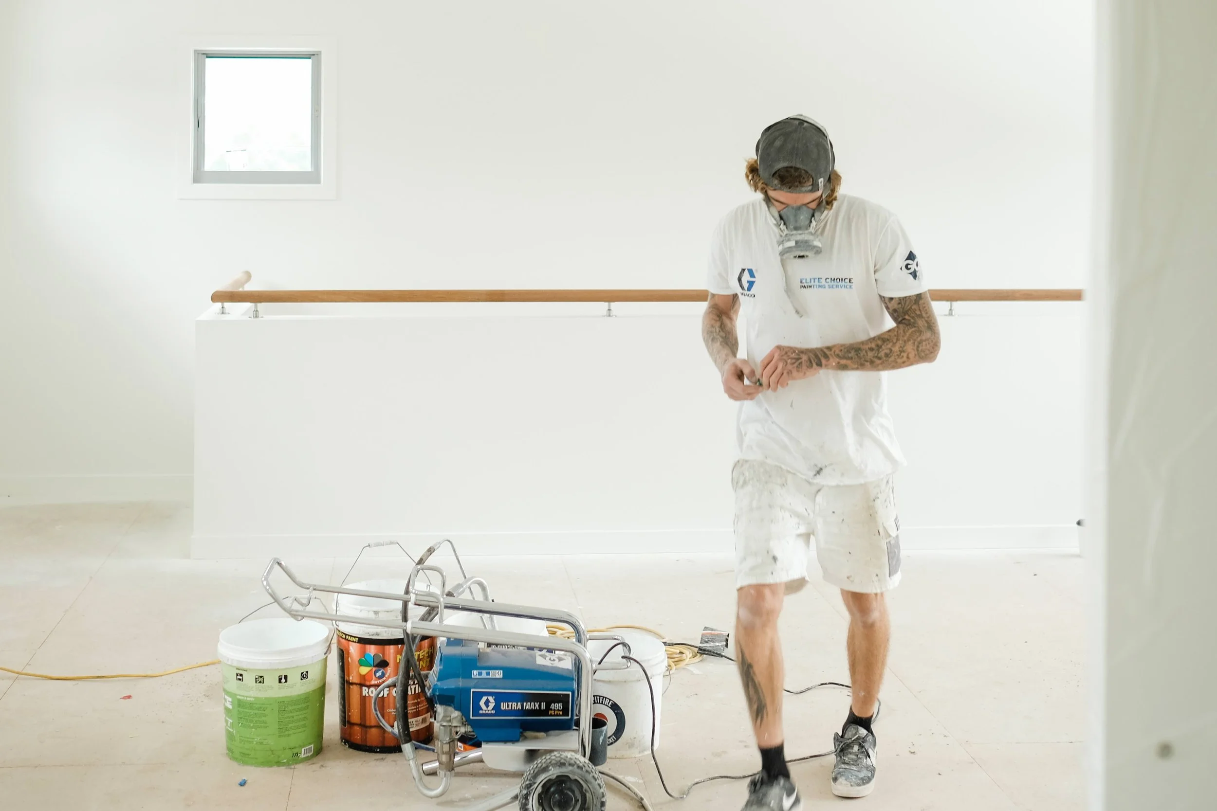 A painter wearing a respirator mask, white shorts, a white T-shirt, and a cap, standing in a bright, white room with a small window, surrounded by painting equipment and supplies.