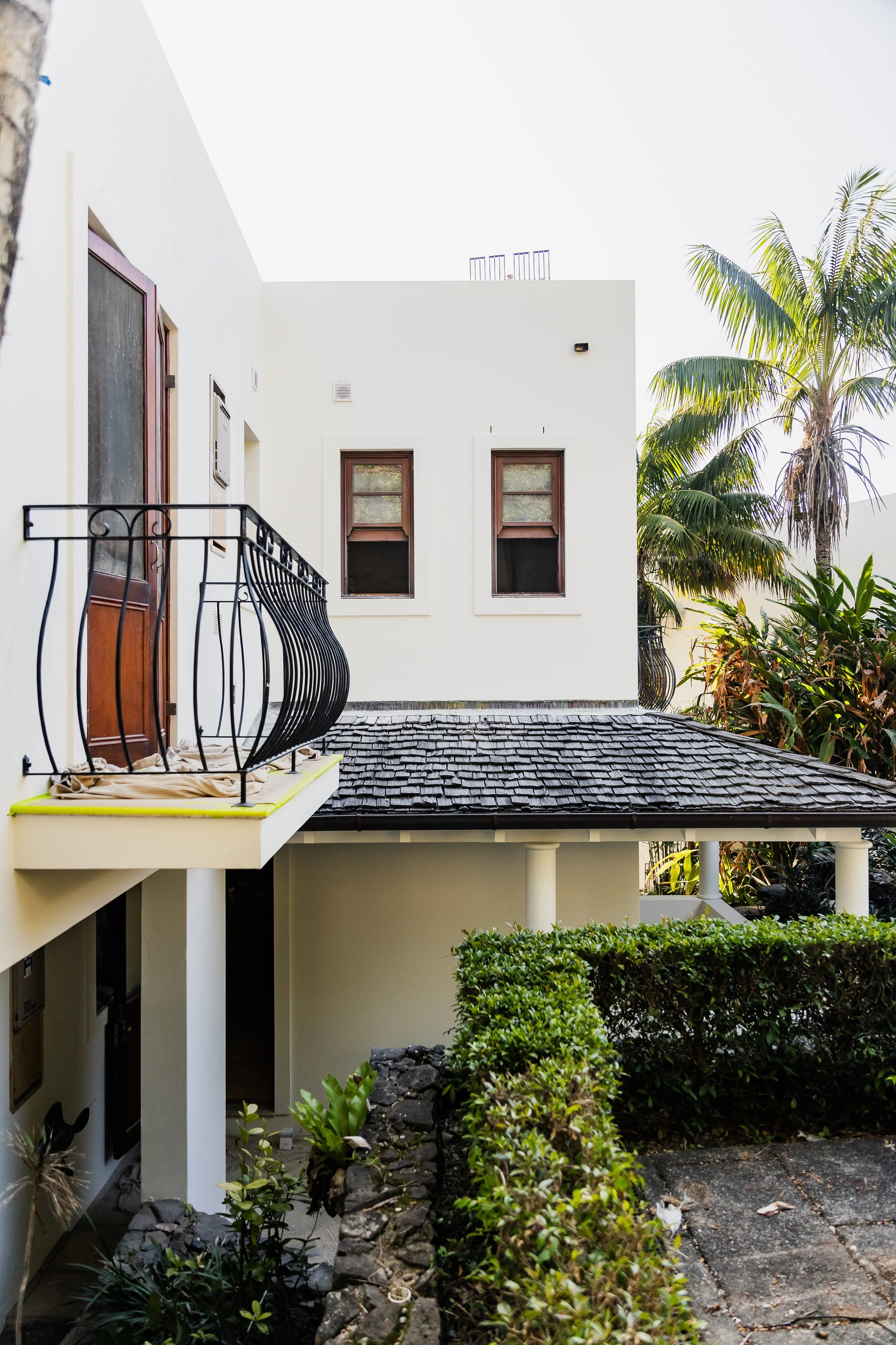 A modern white building with two windows, a small balcony with a black wrought-iron railing, and tropical plants including palm trees and bushes.