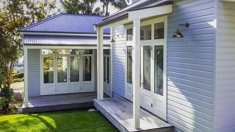 White house with a blue metal roof, featuring a small porch with wooden steps, windows, and a glass door. The house is surrounded by a green lawn and trees.
