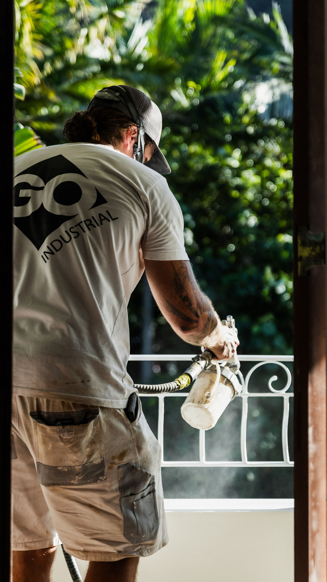 A worker wearing a white G&O Industrial t-shirt, beige shorts, and a cap, is using a spray gun to paint or apply coating on a balcony railing with a lush green background.
