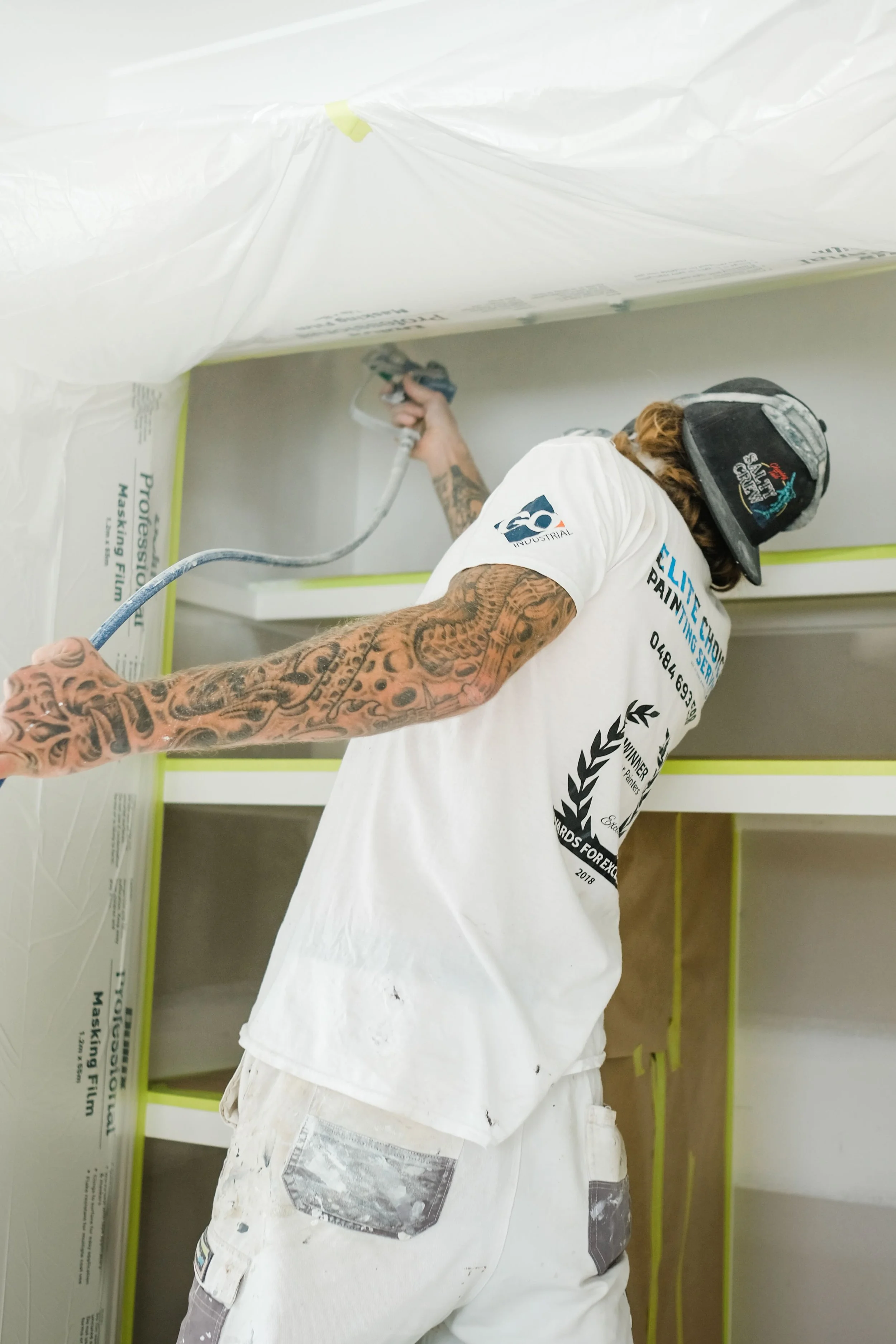 A tattooed construction worker spray-paints a wall inside a room, wearing a helmet and white work clothes.