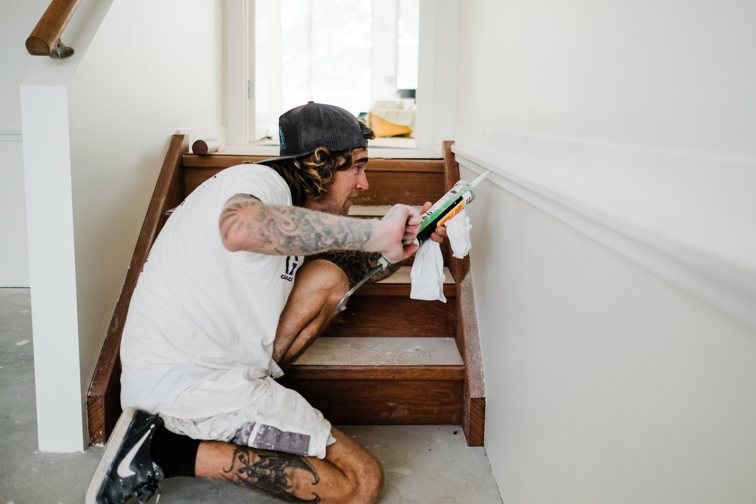 A man with tattoos, wearing a black cap and white shirt, crouches on the floor applying caulk to the bottom of a wooden staircase railing in a light-colored room.