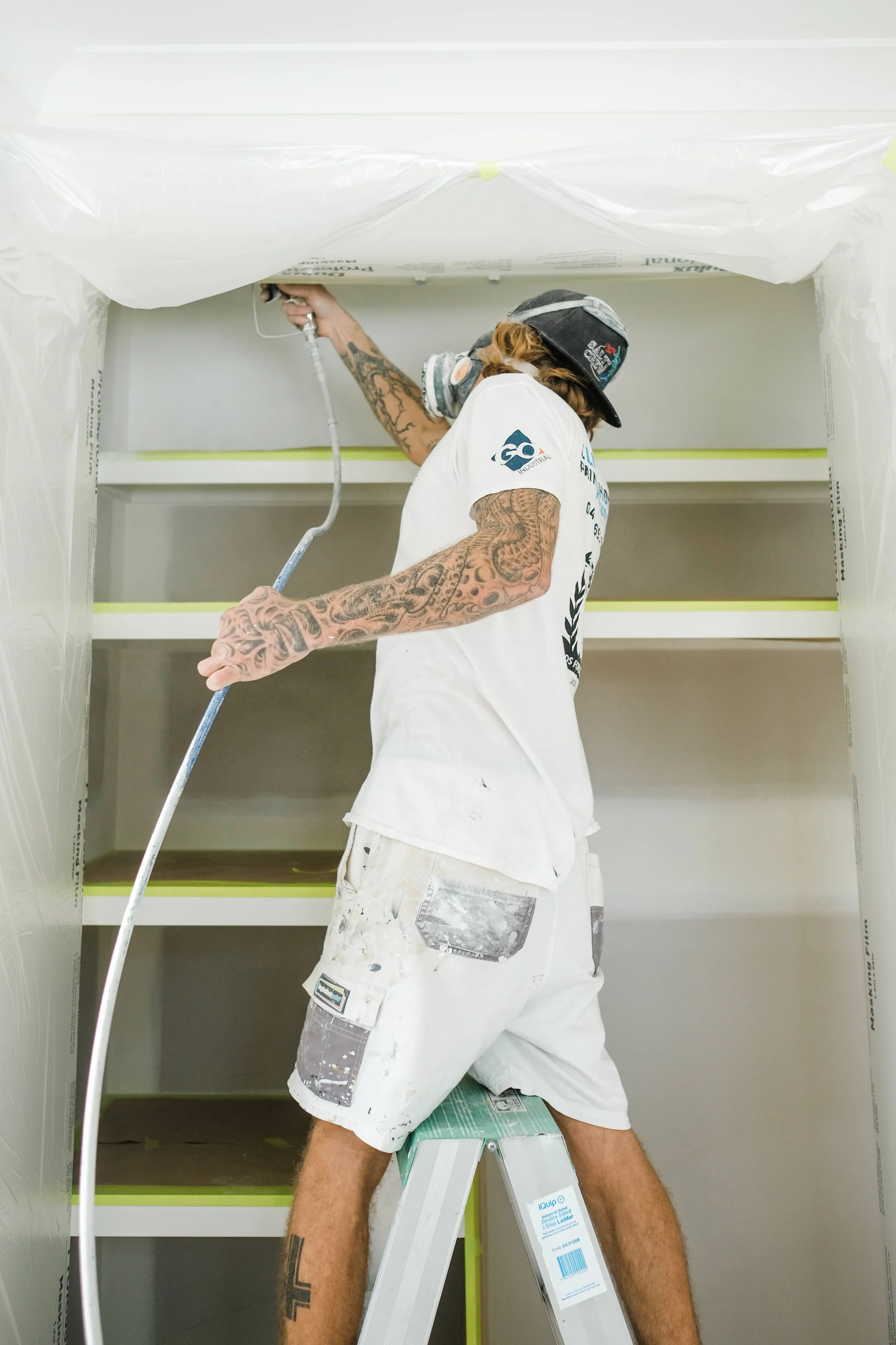 A person with tattoos wearing a white shirt, shorts, and a cap painting the upper shelves of a built-in bookcase with a roller brush.