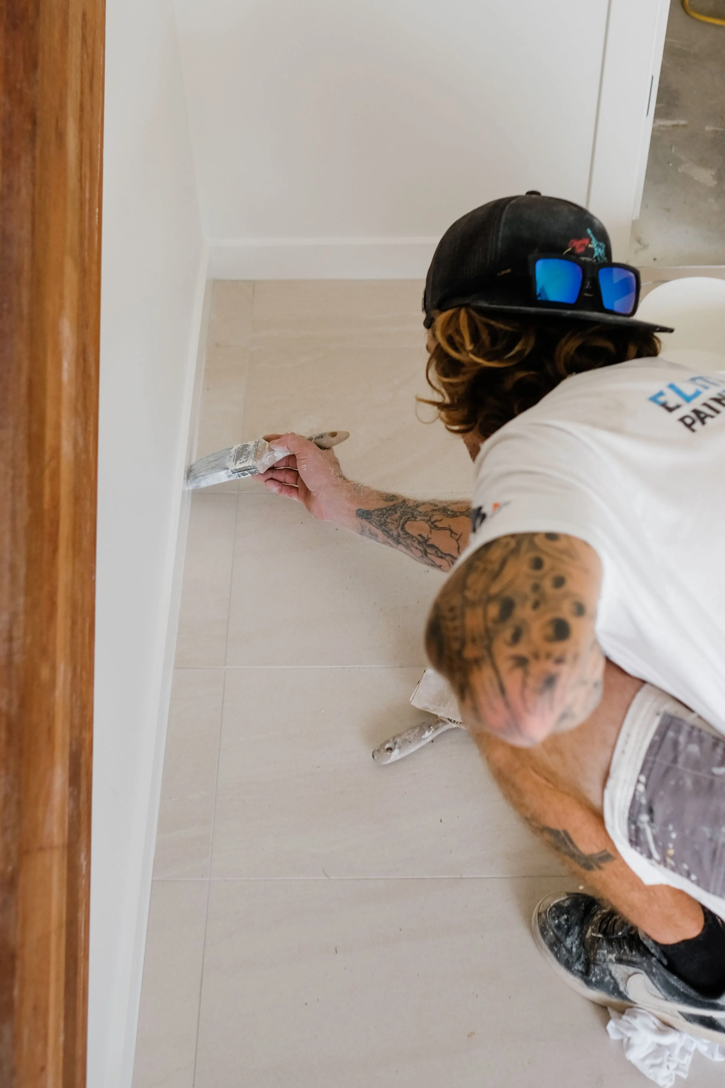 Person painting a beige tiled wall with a paintbrush, wearing a black cap with sunglasses on top, white T-shirt, and showing tattoos on arms.