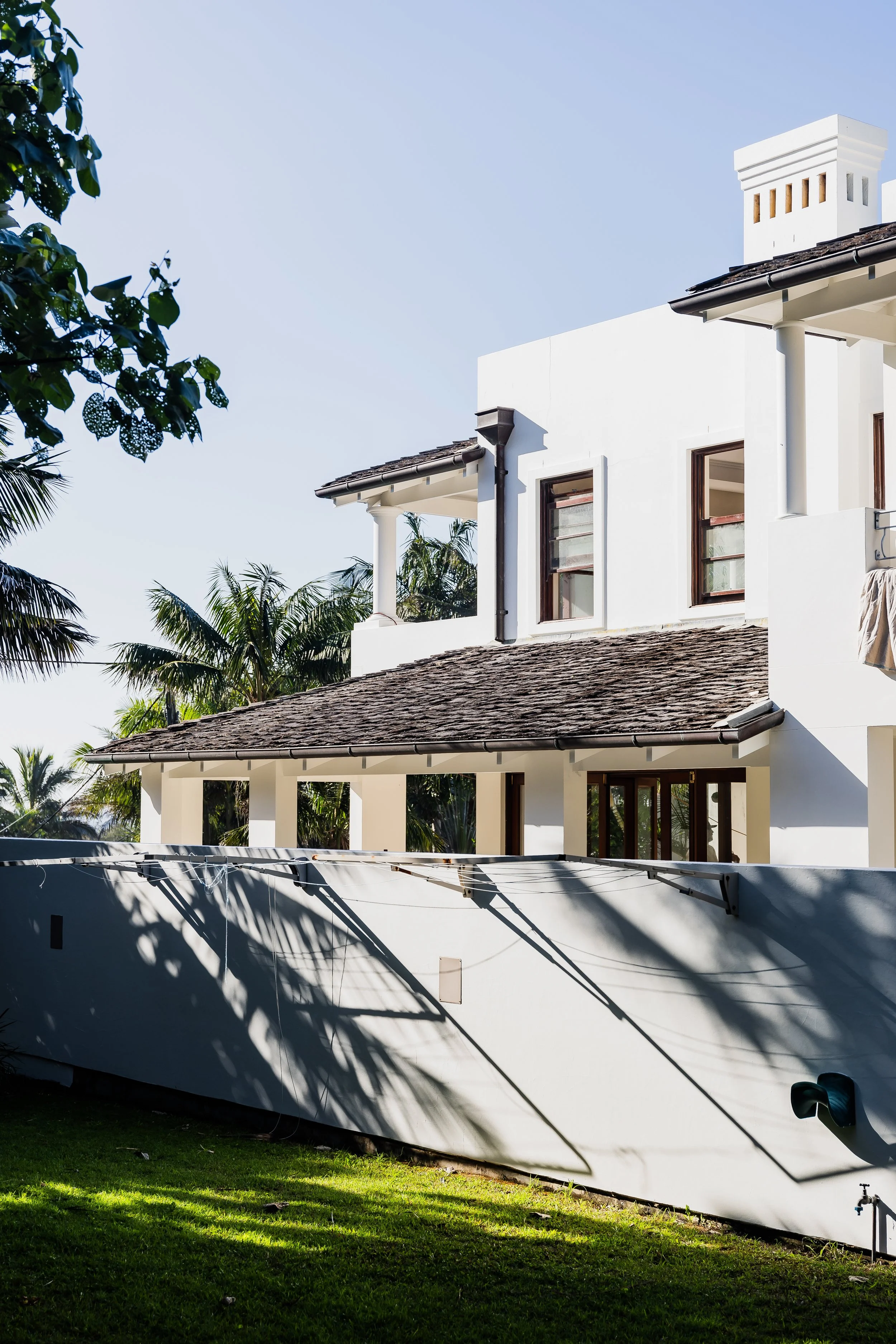 The image shows a white house with a tiled roof, surrounded by greenery and palm trees, with sunlight casting shadows on the exterior wall.