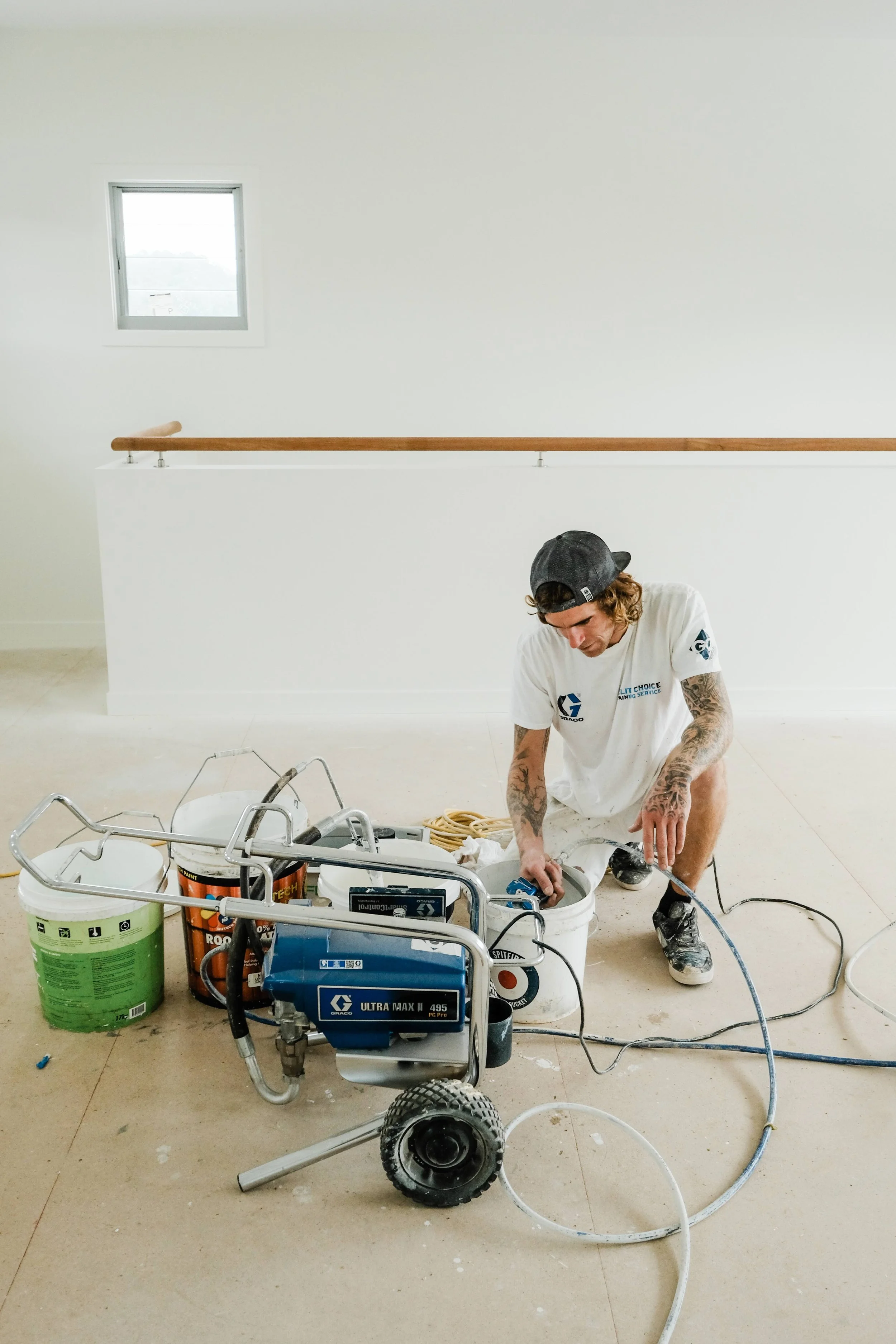 A man kneeling on the floor working with construction equipment, surrounded by buckets and tools, in a room with a window on the wall.
