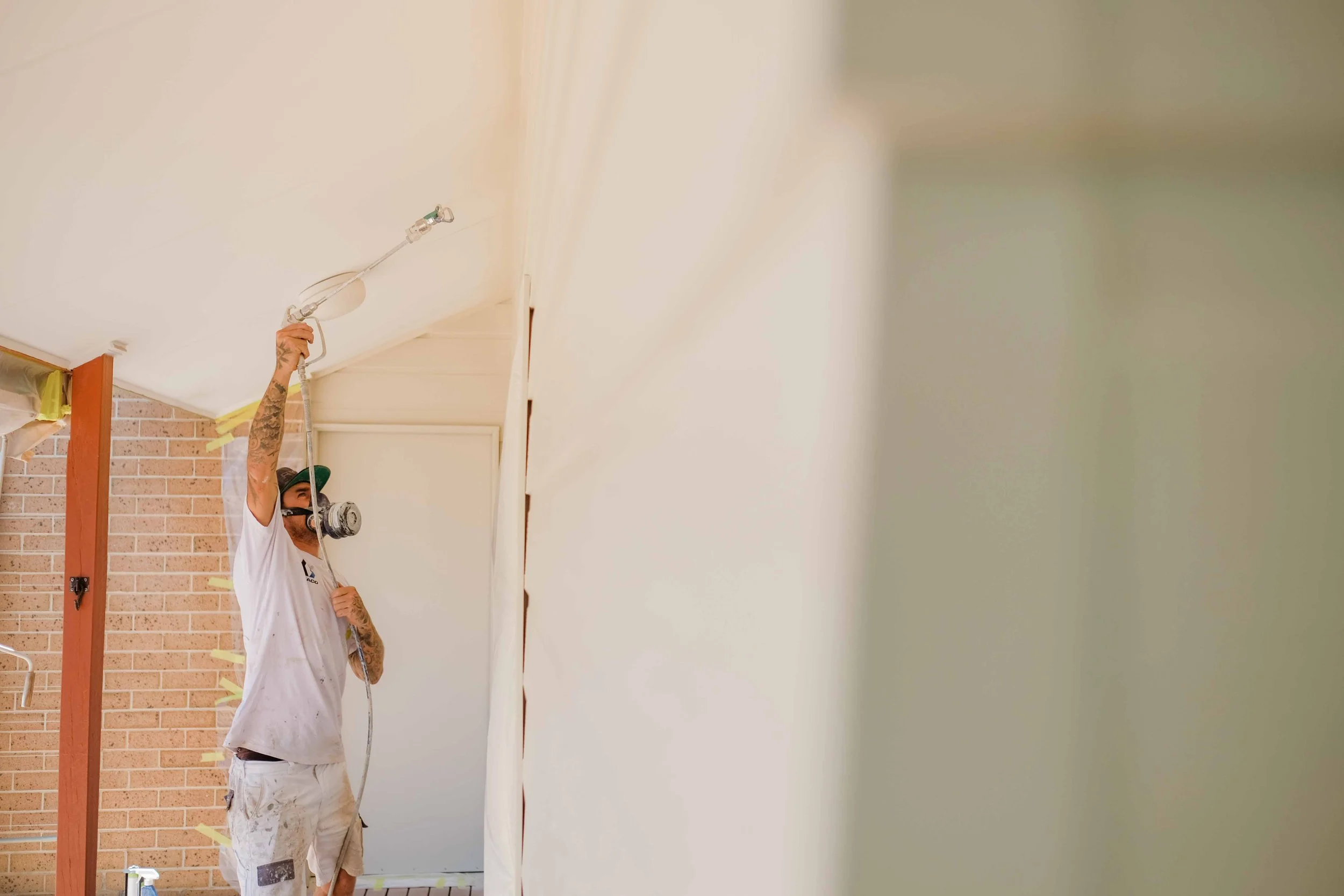 A person wearing a mask and a white shirt with paint stains is spray painting a wall in a room with brick and drywall, standing on a ladder.
