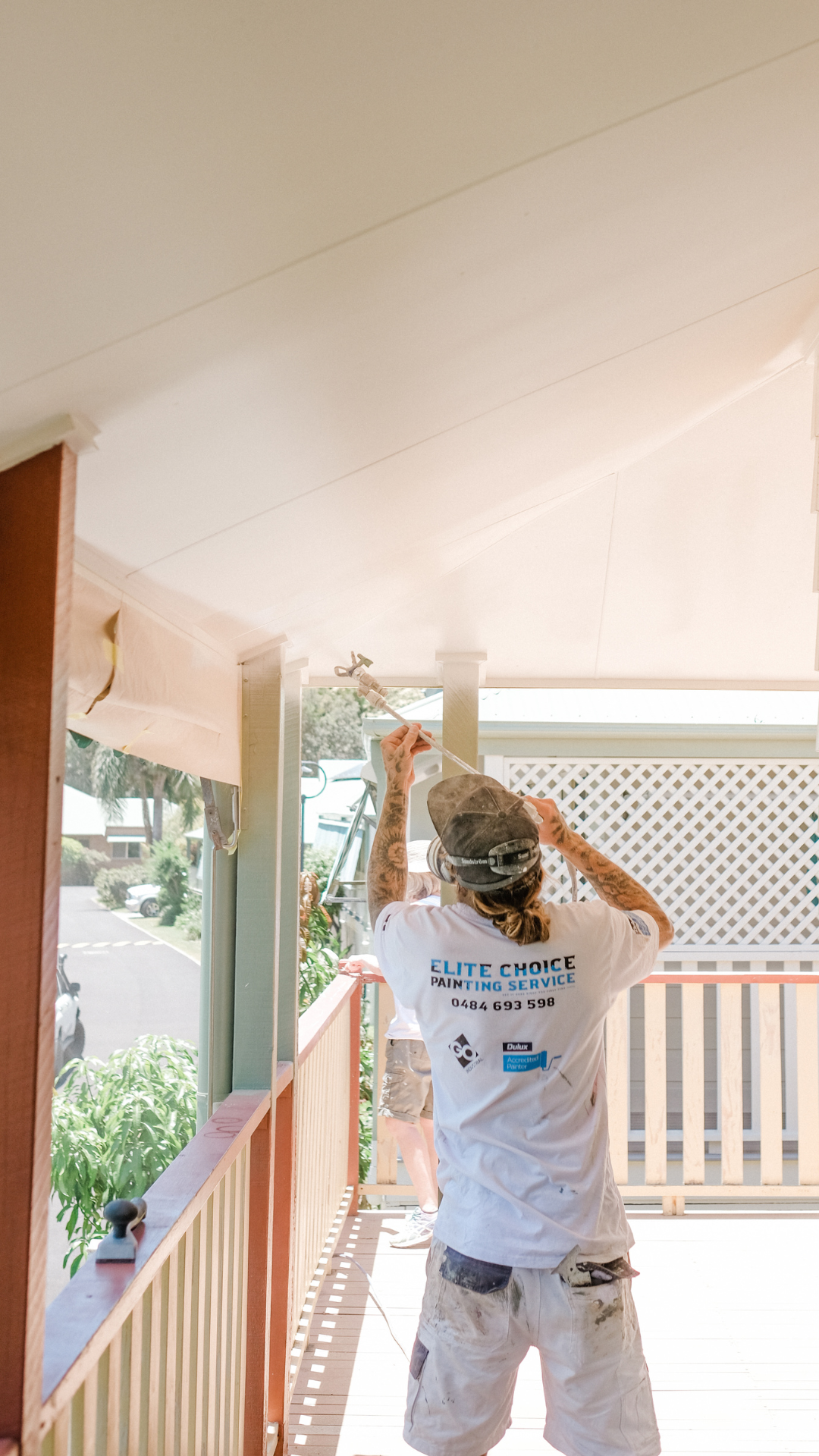 A painter from Elite Choice Painting Service spraying paint on a ceiling. The painter is wearing a gray cap, white T-shirt, and white shorts, standing on a wooden deck.
