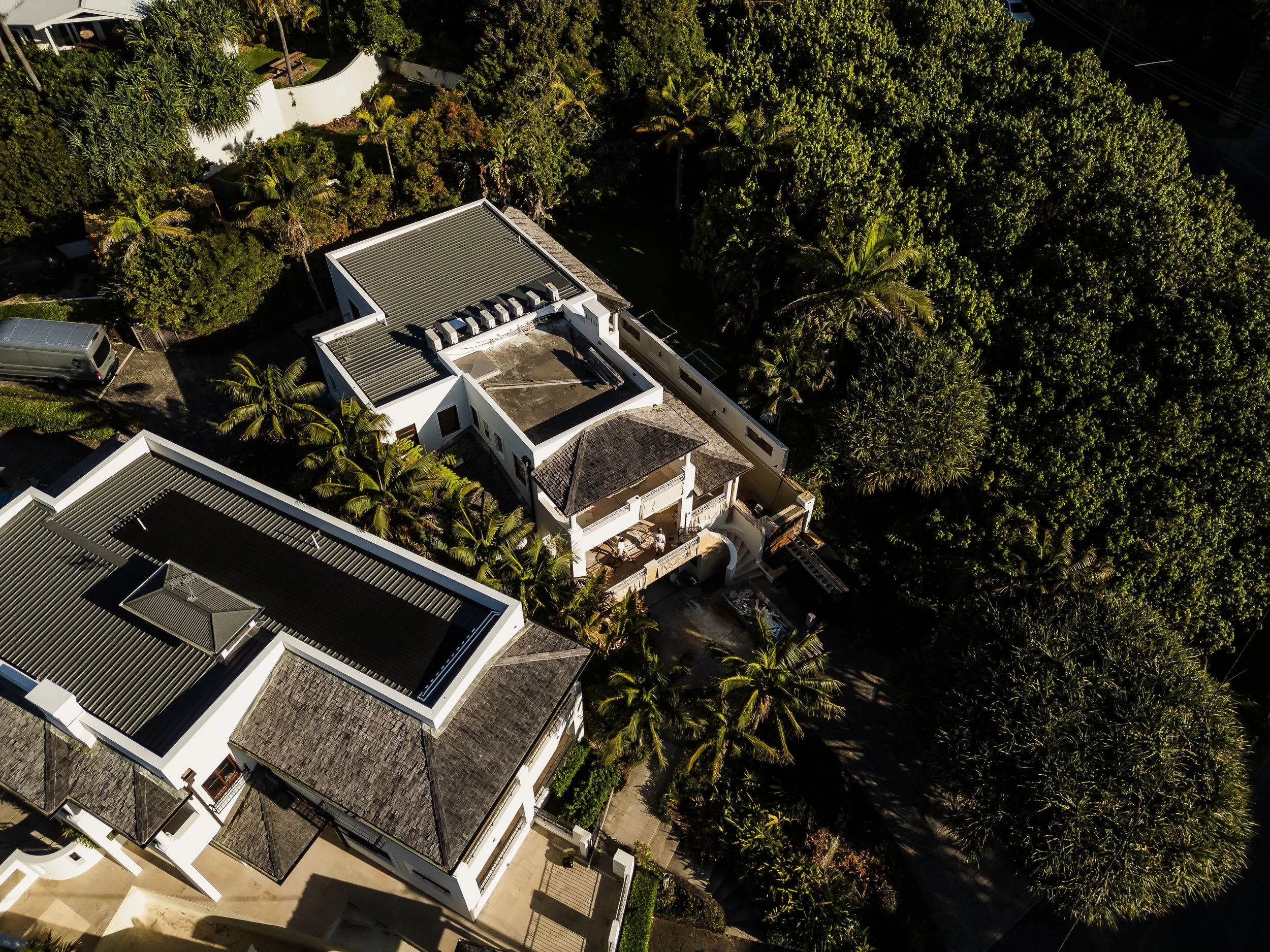 Aerial view of a modern white house with multiple rooftops surrounded by dense green trees and a driveway with parked cars.
