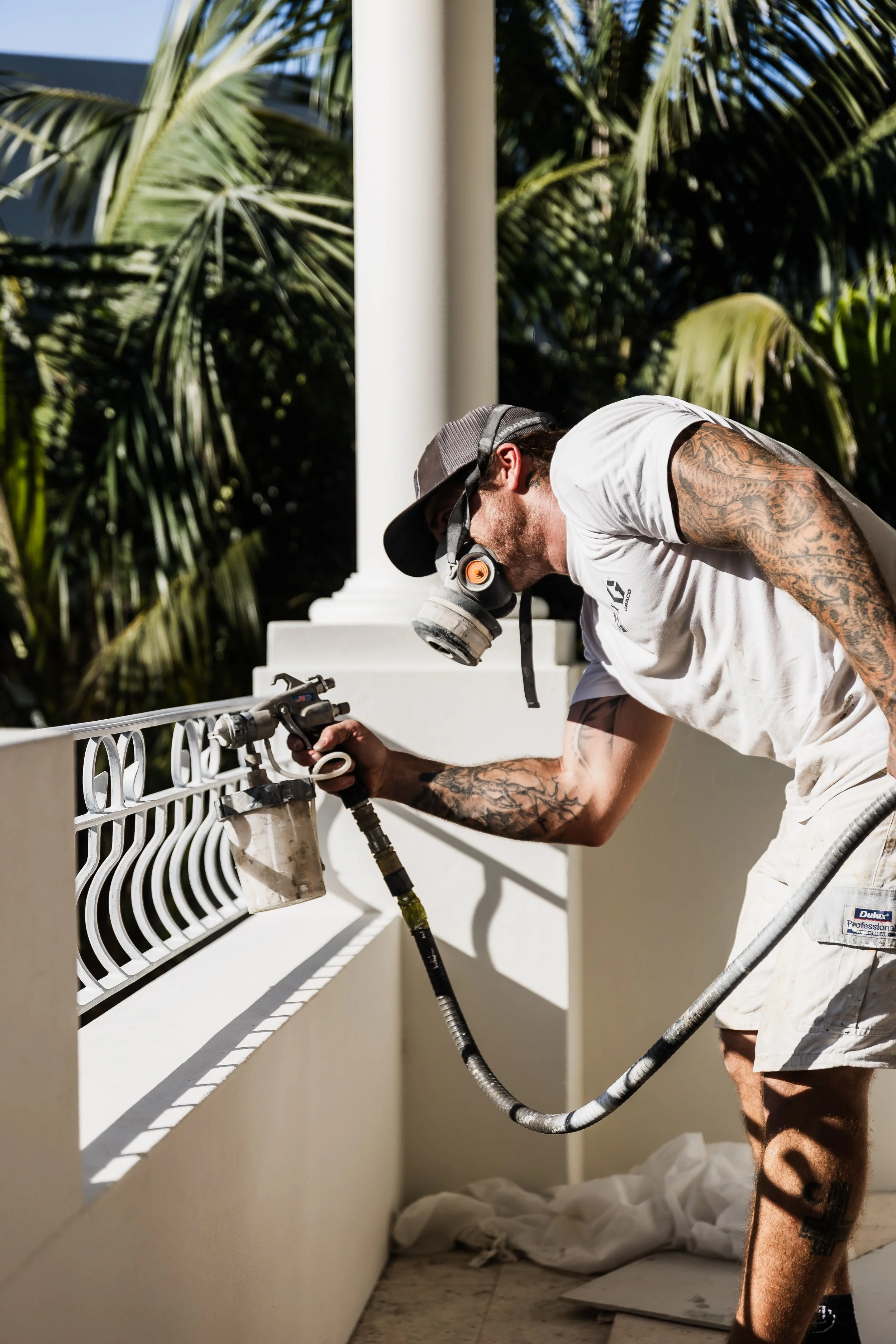 A construction worker wearing a mask and a cap, with tattoos on his arms and leg, is spray painting a metal railing on a balcony.