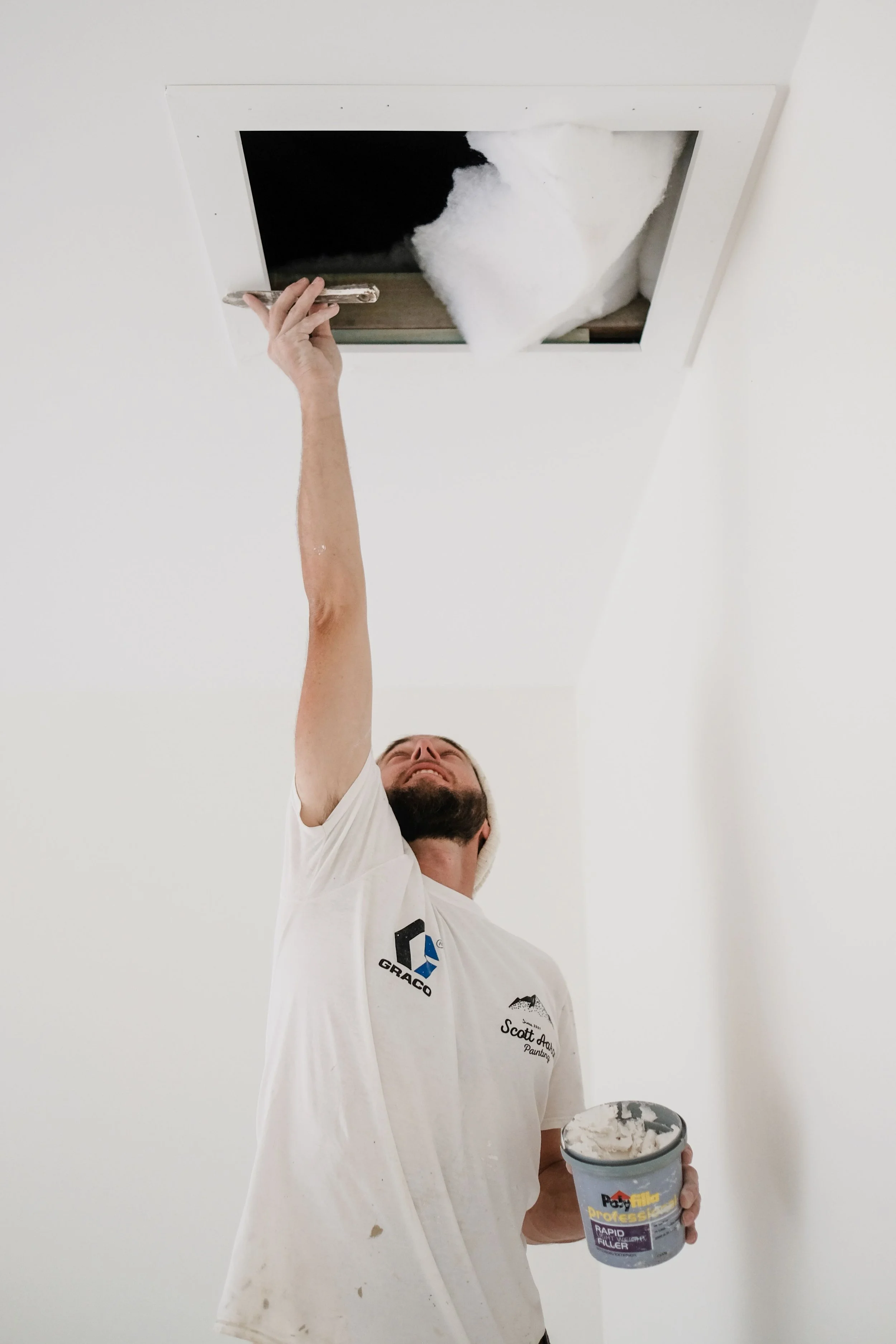 A man wearing a white t-shirt and cap is reaching up to access a ceiling housing with insulation. He is holding a container of joint compound.