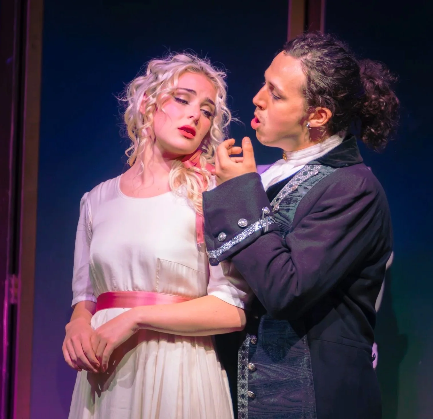Two women in a theatrical scene, one with curly blonde hair and a white dress, the other with dark curly hair in a dark coat, engaged in an intense conversation.