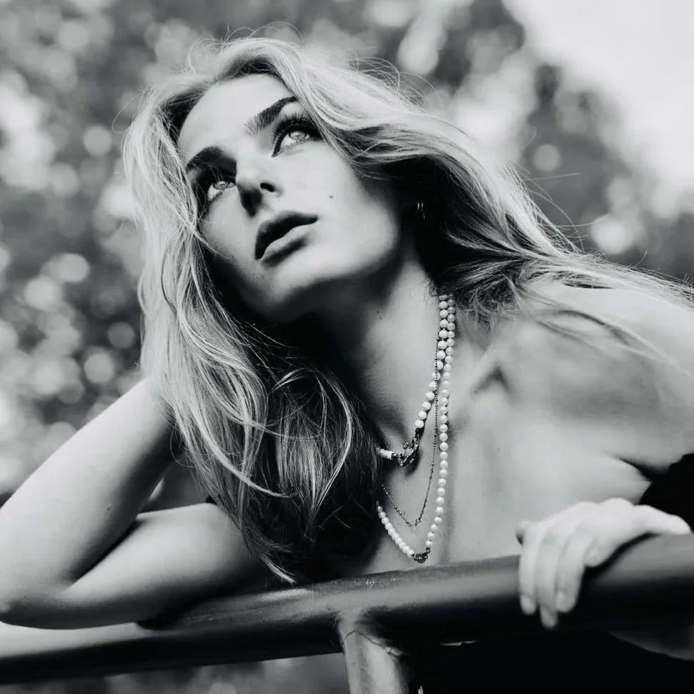 A black and white portrait of a woman with wavy hair and layered necklaces, looking upward while resting her head on her hand outdoors.