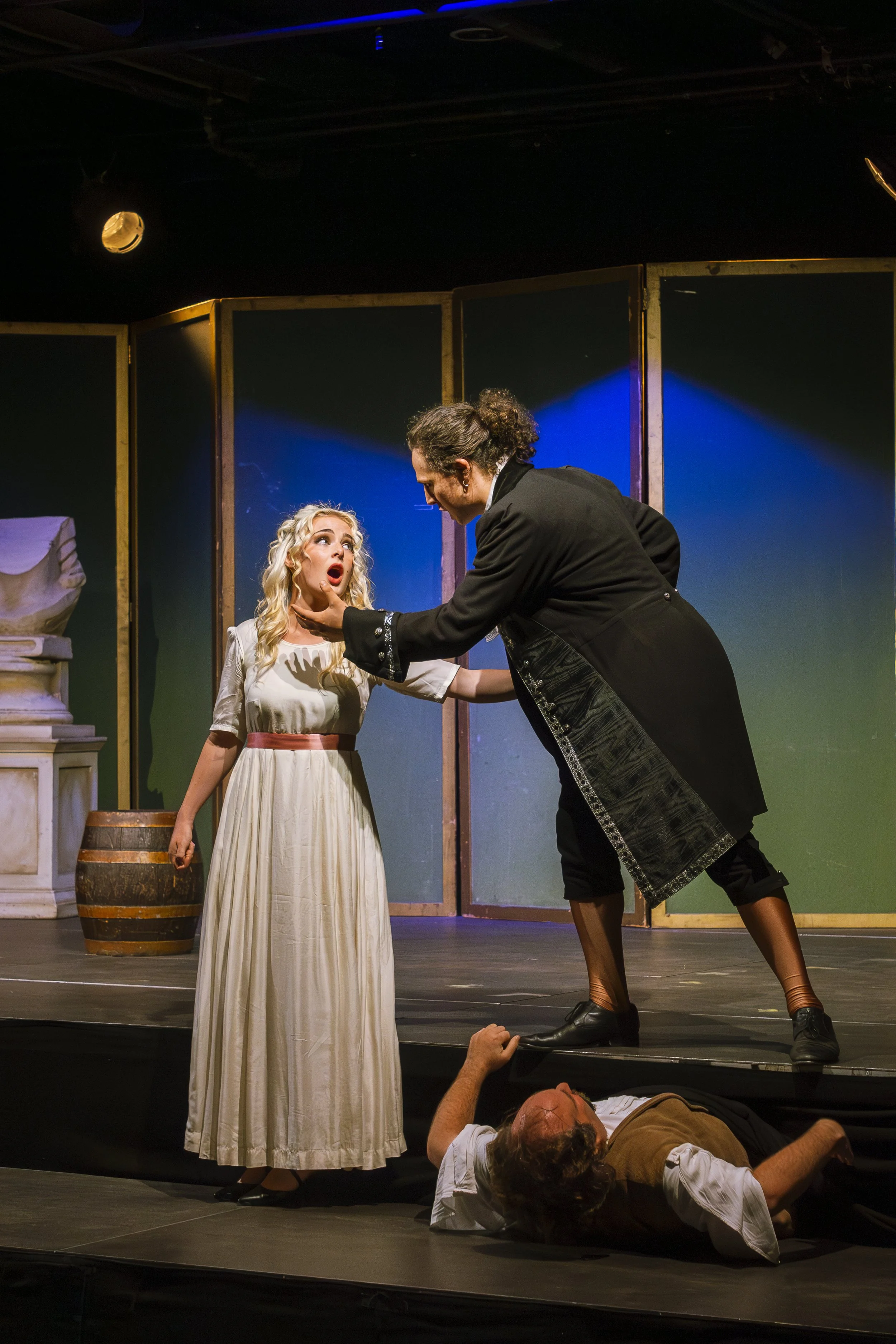 Actors perform a dramatic scene on stage, with one man holding the chin of a woman dressed in a long white dress, while another man lies on the ground beneath them.
