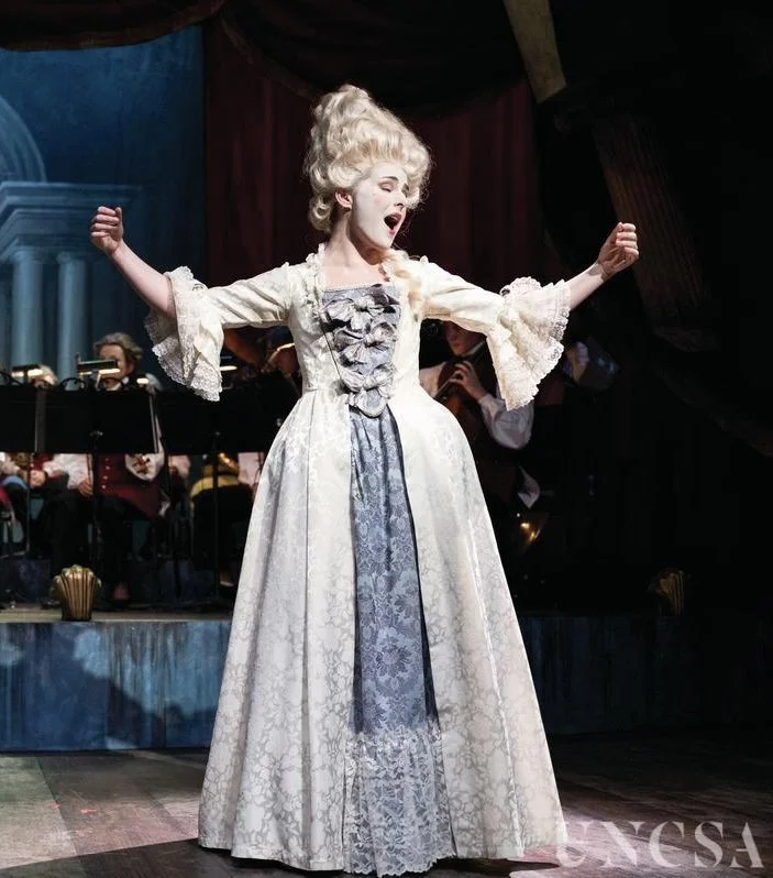 Woman dressed in period costume on stage, singing with arms raised, with orchestra in the background.