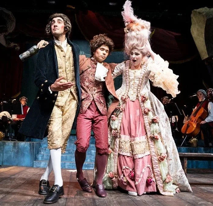 Three actors dressed in elaborate 18th-century costumes standing on stage, with musicians playing in the background.