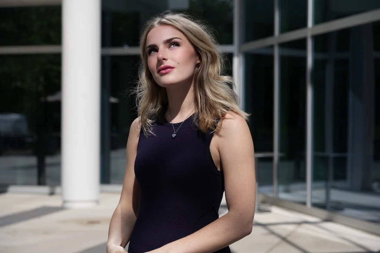 A young woman with blonde hair and fair skin wearing a sleeveless black dress, standing outdoors near a modern glass building.