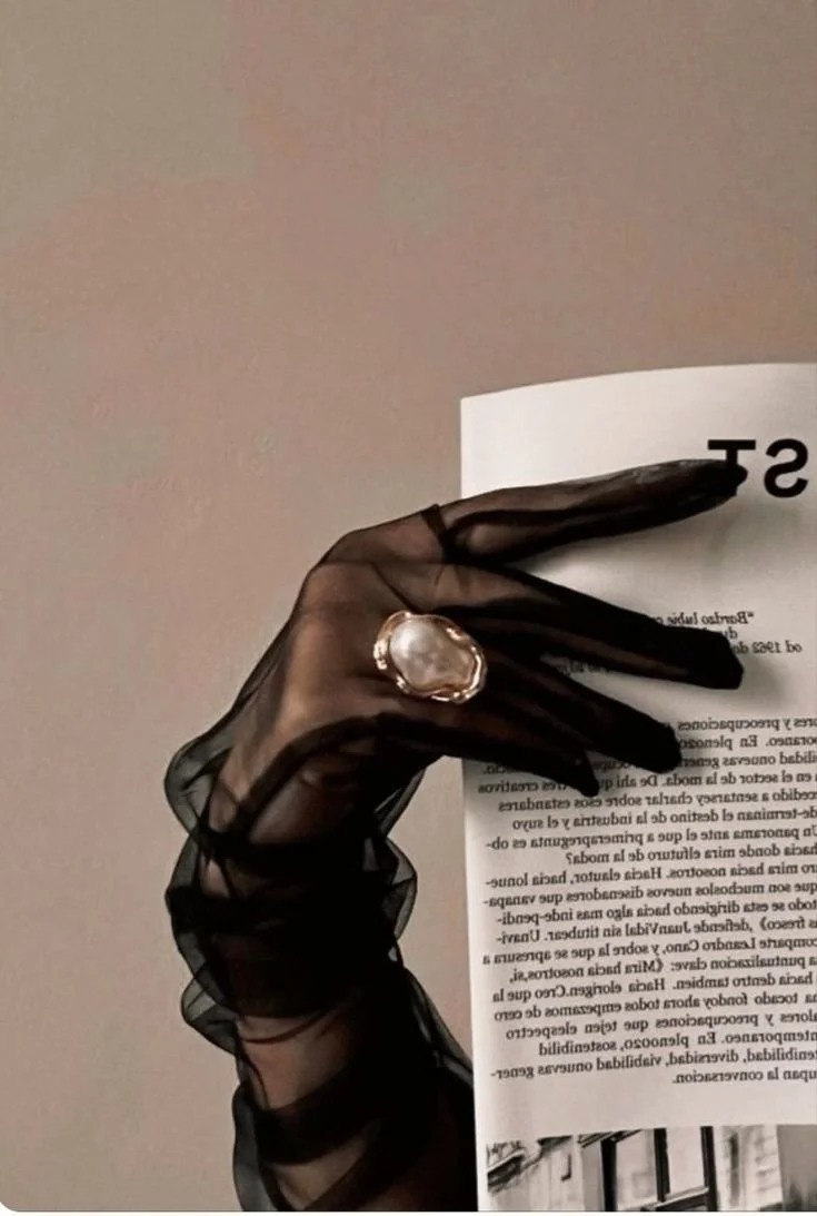A black sheer glove with a large pearl ring worn on the middle finger, gripping a piece of paper with printed text.