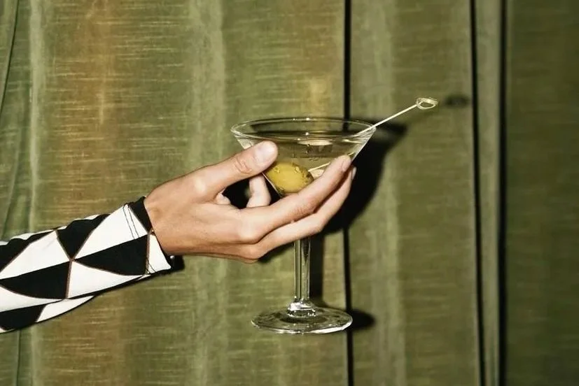Person holding a martini glass with olives inside and a cocktail pick resting on the rim, against a wooden background.