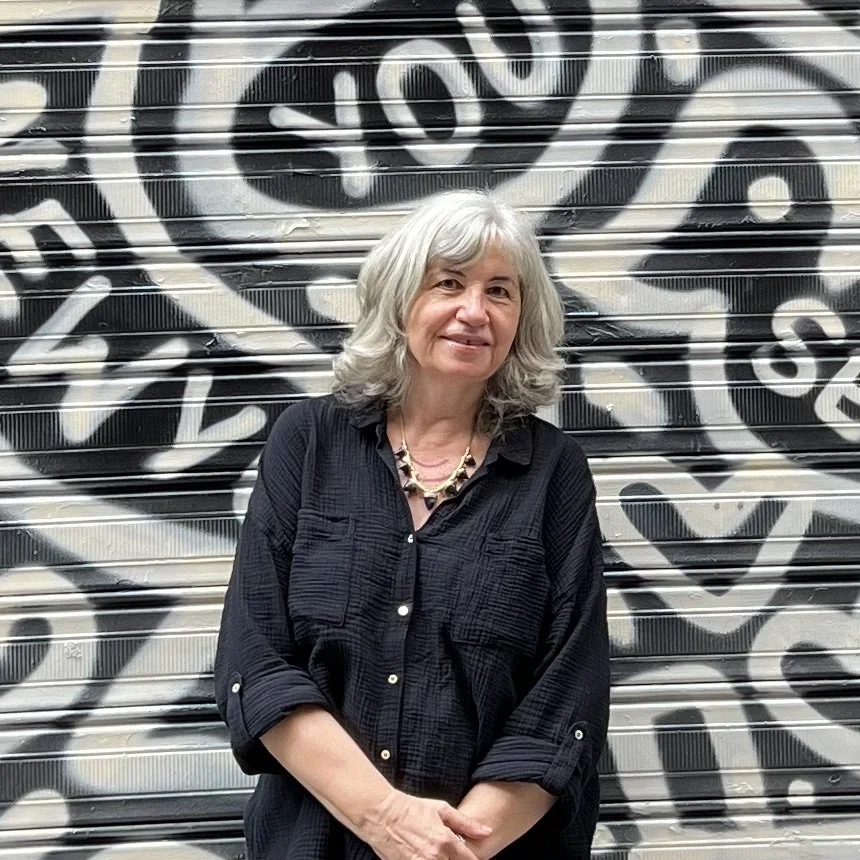 A woman with gray, wavy hair standing in front of a graffiti-covered metal shutter door.