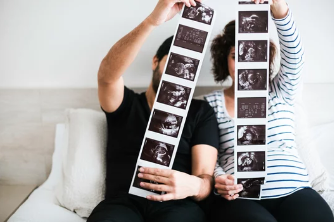 Two women sitting on a bed holding ultrasound photo strips in front of their faces, showing different ultrasound images.