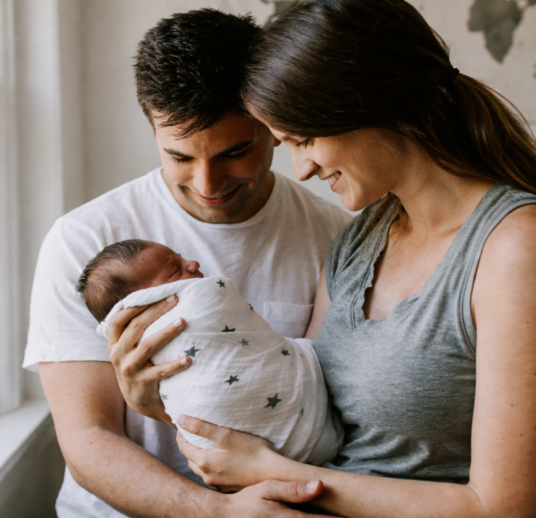 Mum and dad hold new born baby who is wrapped in a muslin blanket and is fast asleep mum and dad look lovingly at their new baby