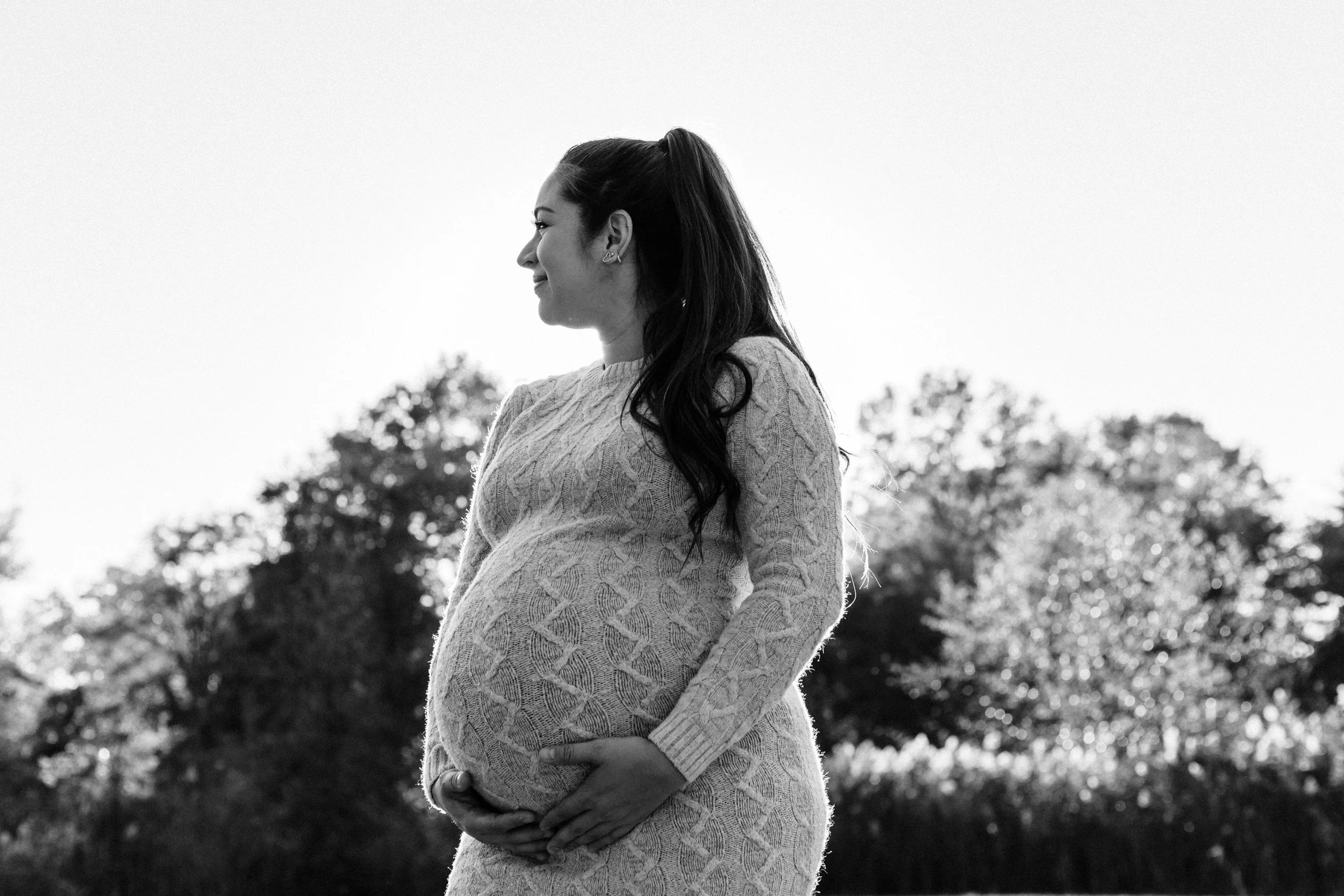 Black and white photo of a pregnant woman outdoors, wearing a knit sweater, with her hands on her belly, smiling and looking to the side, with trees in the background.